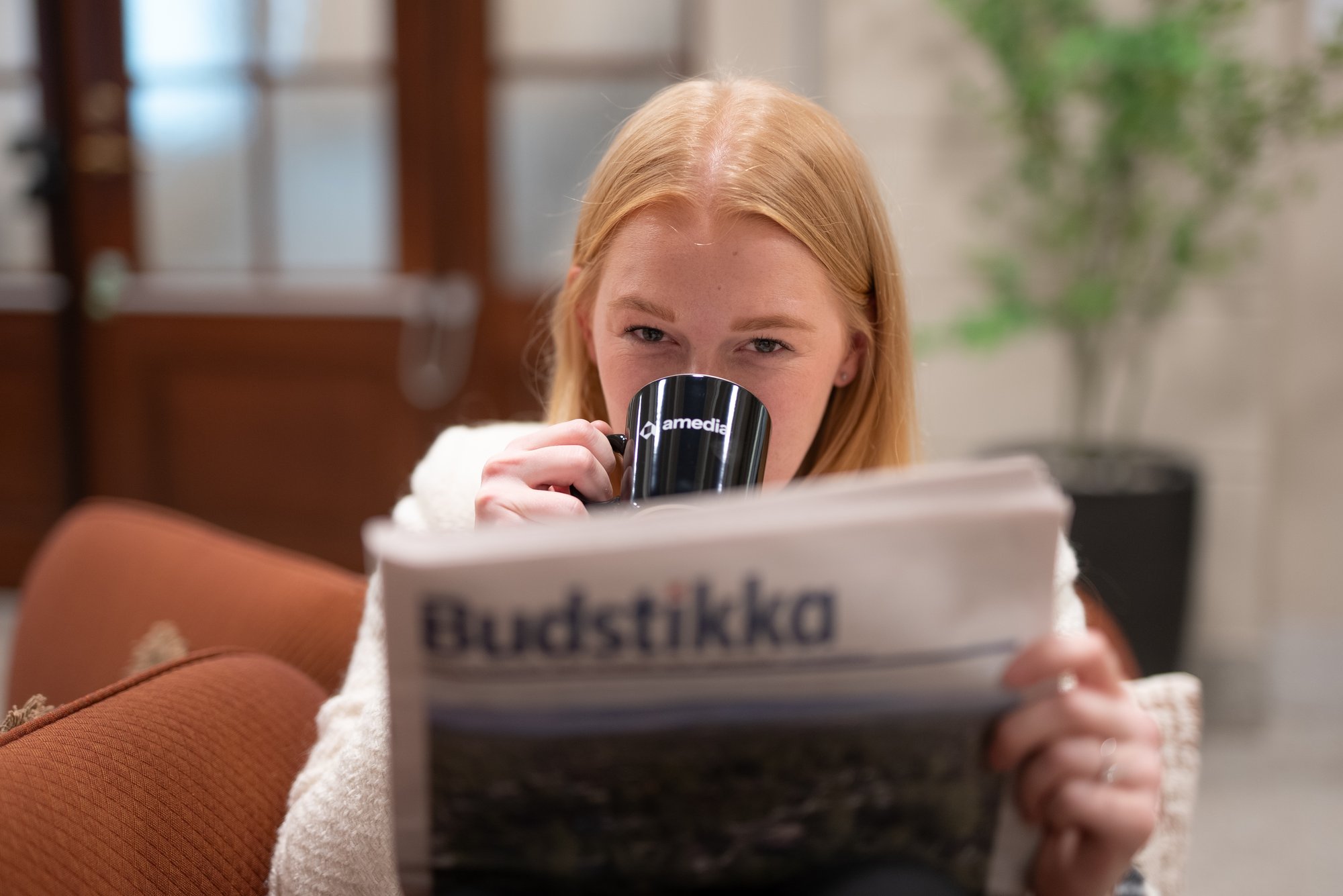 Kvinne drikker kaffe av Amedia-koppen mens hun leser Budstikka og ser lurt inn i kamera - Amedia Annonse